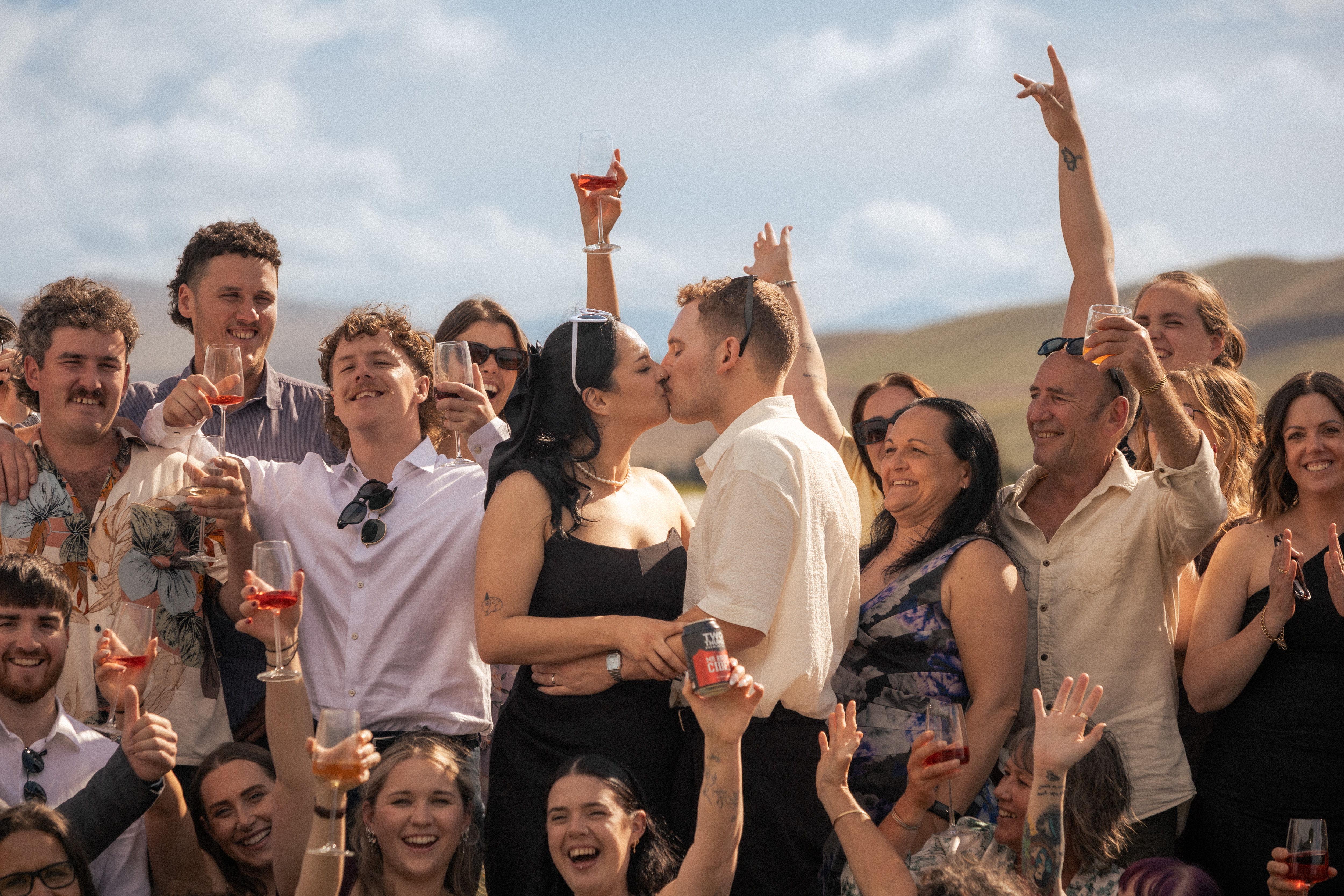Newlyweds kissing while surrounded by cheering wedding guests raising their glasses in the hills