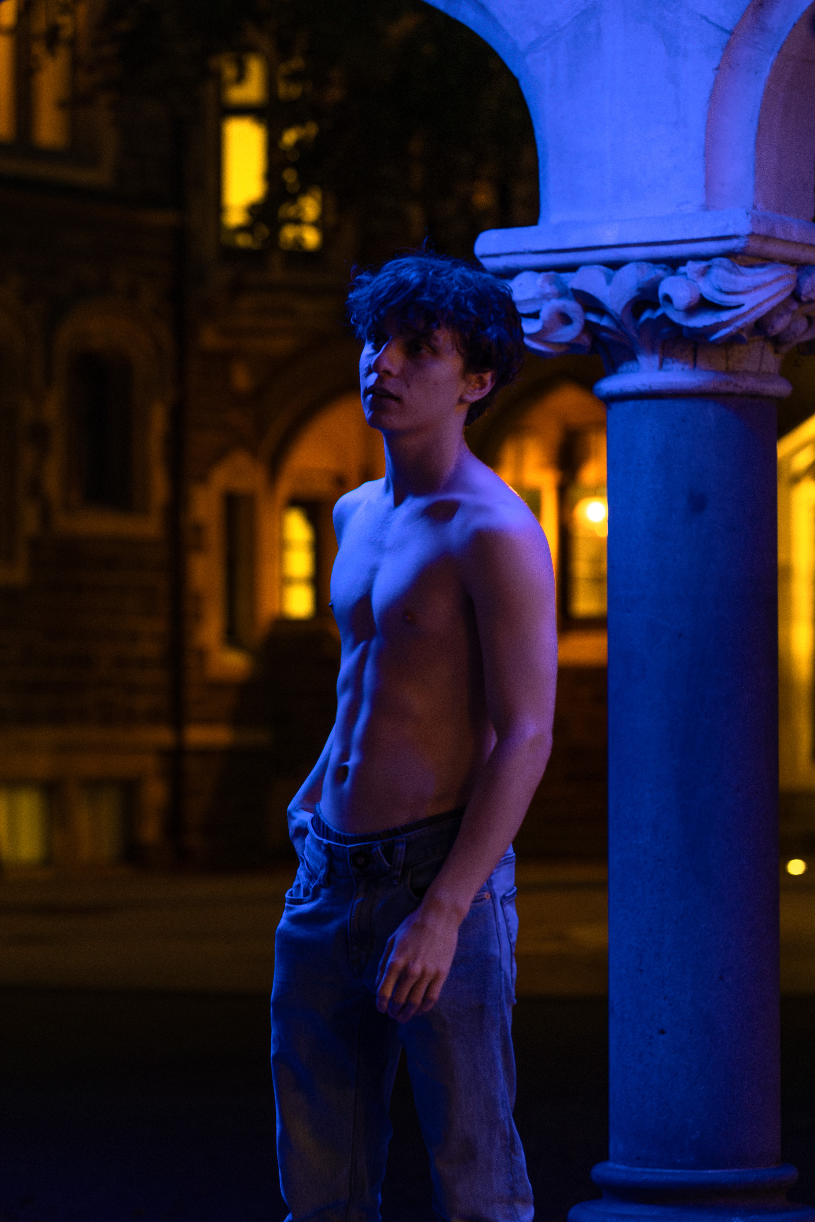 Performer standing beside a blue-lit stone column at night, city windows glowing behind