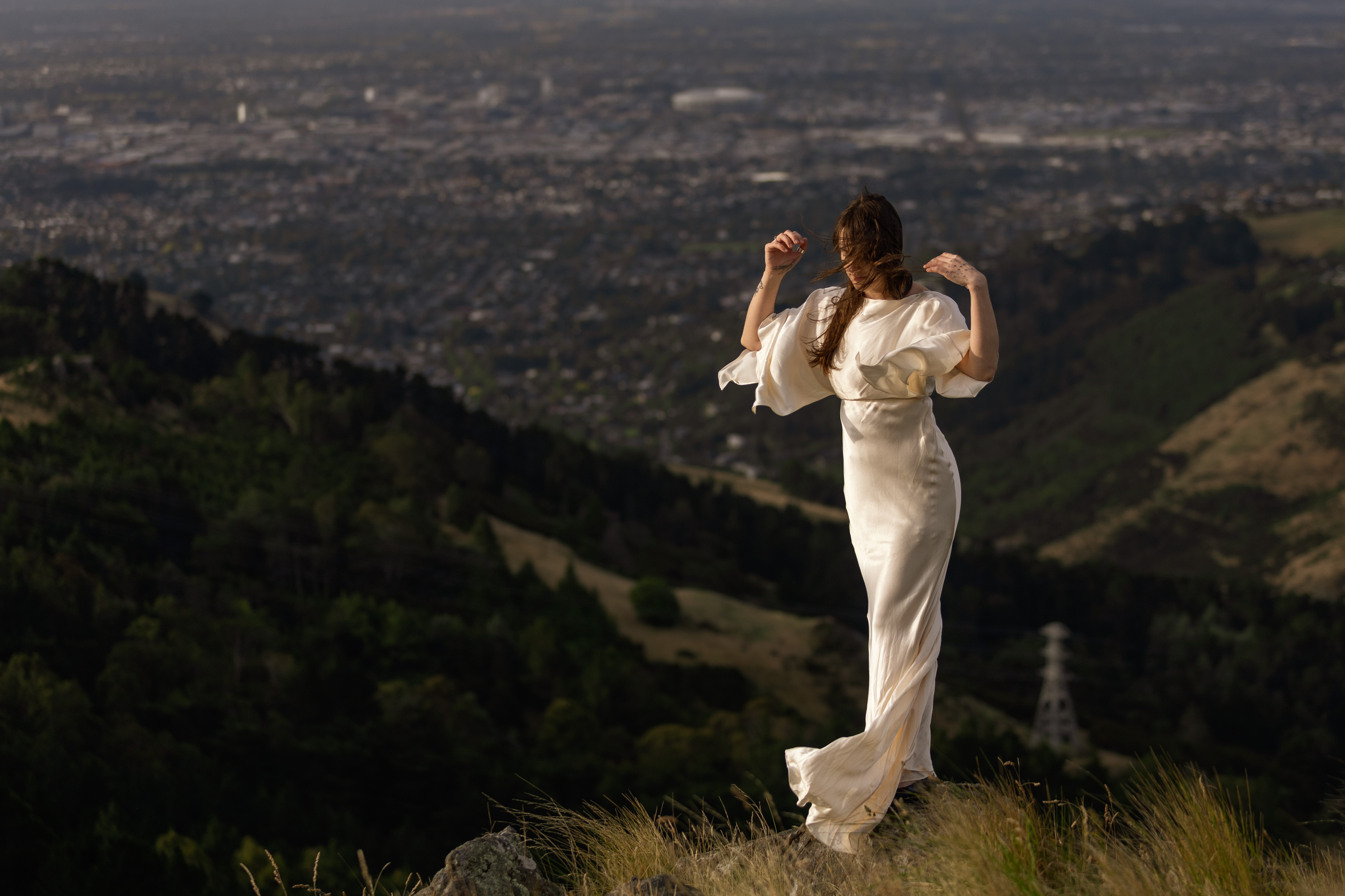 Woman in a flowing white dress standing on a hilltop overlooking the city below
