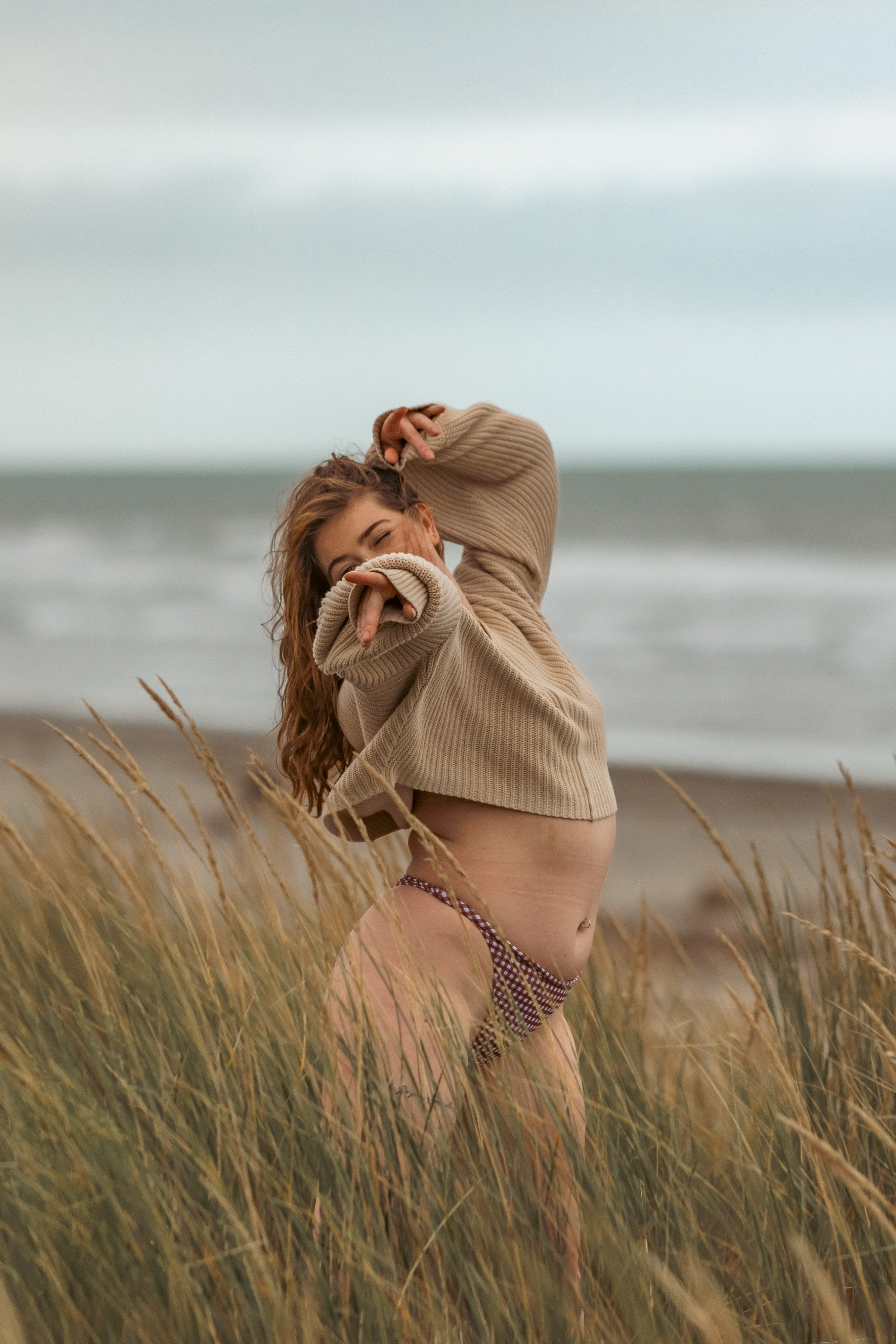 Woman in a cream jumper standing in tall dune grass by the sea, arm raised through her hair